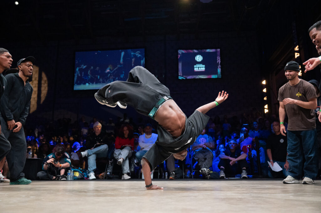 un danseur de breakdance fait une figure tête à l'envers avec seulement la main sur la scène avec en fond tout le public réunis autour de celle-ci dans une salle sombre
