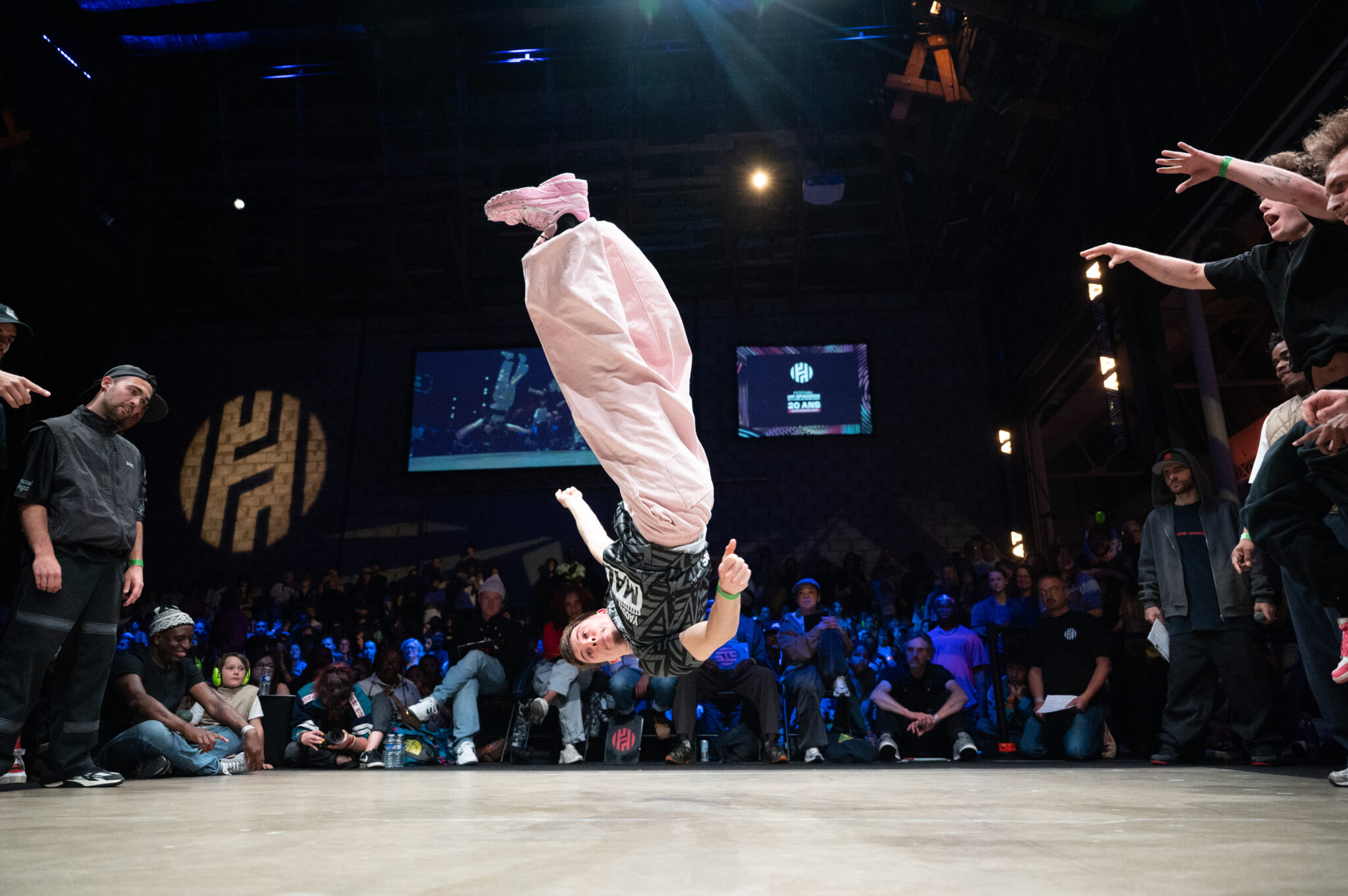 un danseur de breakdance fait une figure en l'air tête à l'envers à la verticale sans toucher la scène avec en fond tout le public réunis autour de celle-ci dans une salle sombre