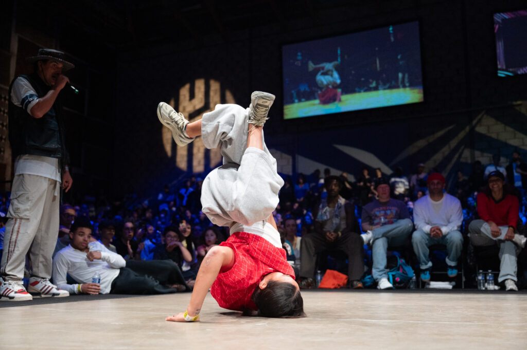 une danseuse de breakdance fait une figure à la verticale sur le sol de la scène sur fond de salle avec de la foule autour