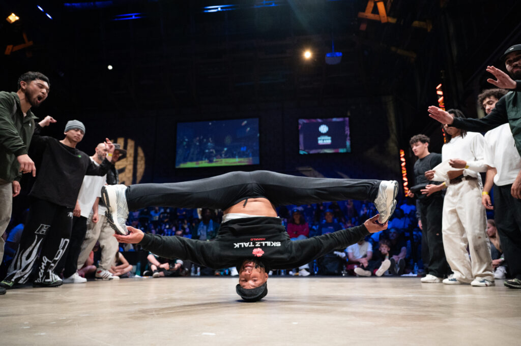 un danseur de breakdance fait une figure tête à l'envers, jambes en grand écart et les mains touchant ses pointes de pied avec seulement la tête sur la scène avec en fond tout le public réunis autour de celle-ci dans une salle sombre