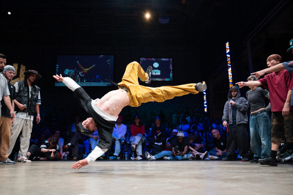 un danseur de breakdance fait une figure à l'horizontale avec seulement la main sur la scène avec en fond tout le public réunis autour de celle-ci dans une salle sombre
