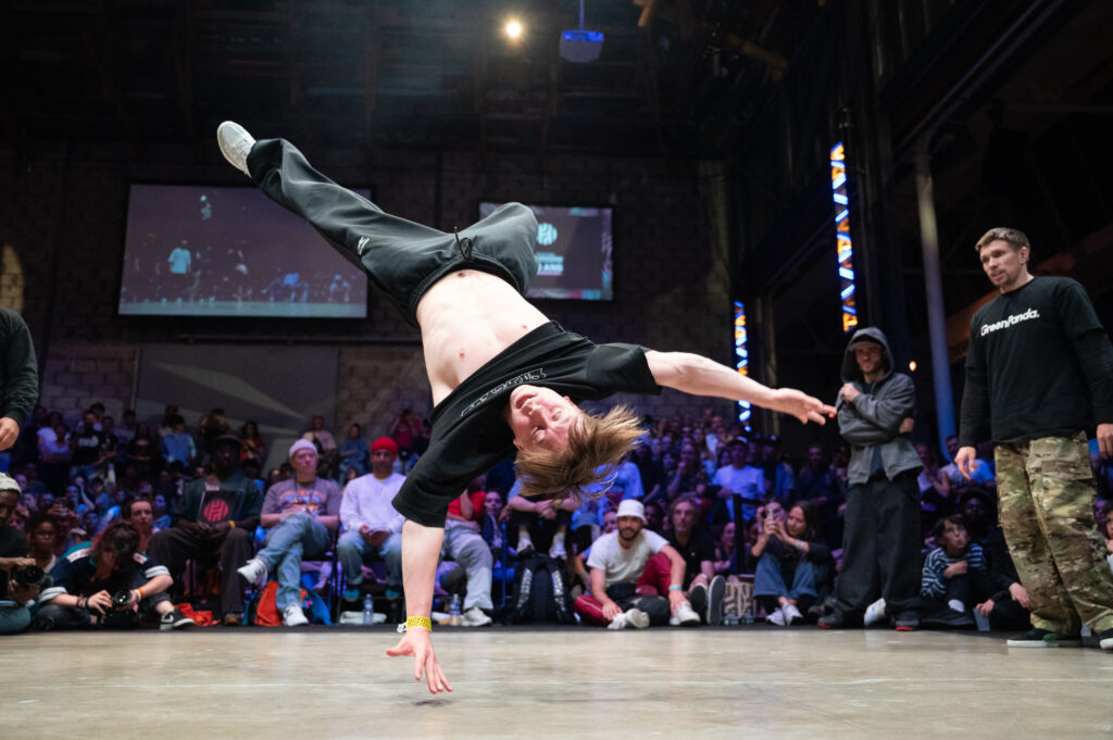 un danseur de breakdance fait une figure tête à l'envers avec seulement la main sur la scène avec en fond tout le public réunis autour de celle-ci dans une salle sombre