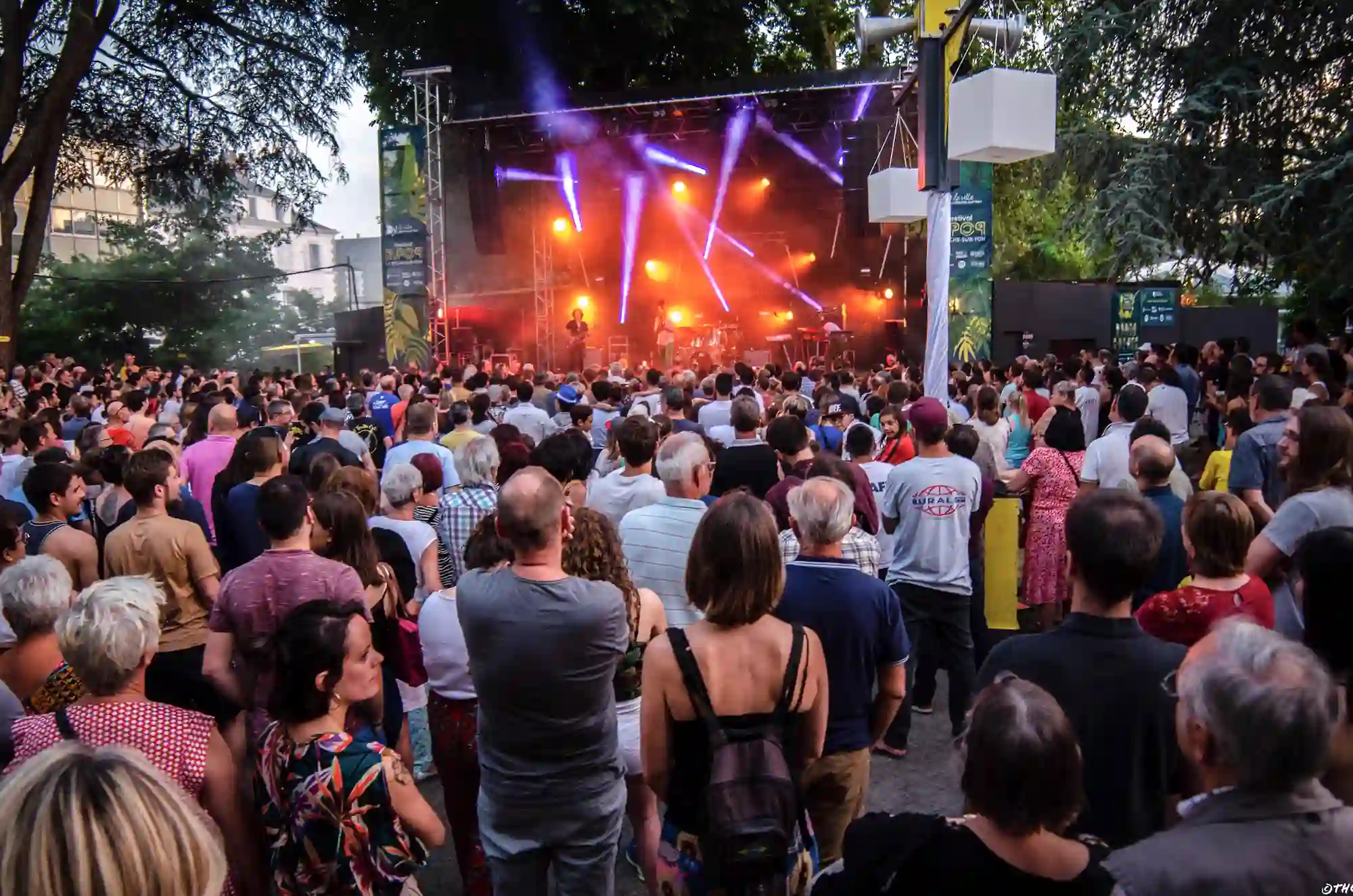 Une foule nombreuse assiste à un concert en plein air en début de soirée. Le public, de tous âges, est tourné vers une scène illuminée par des projecteurs orange et violets. Plusieurs musiciens jouent sur scène, entourés d’arbres et de structures urbaines.