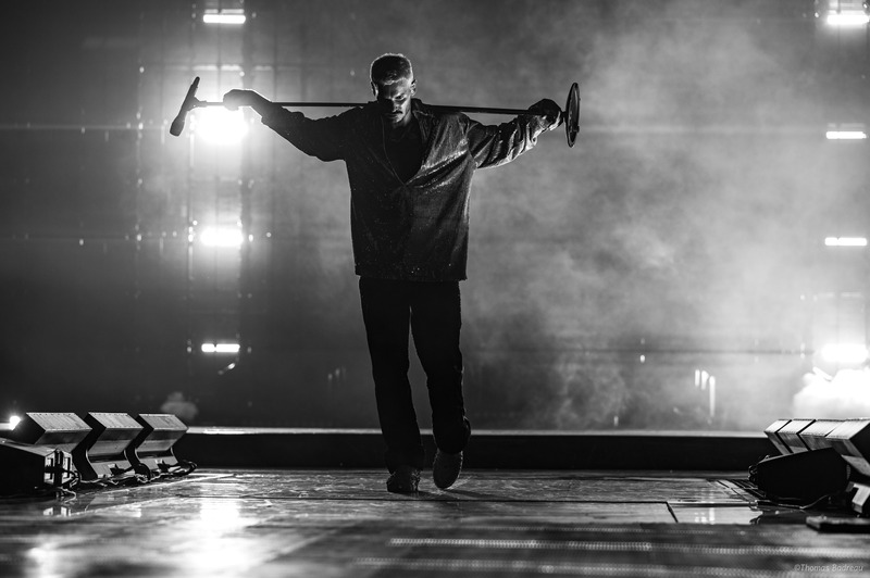 Matt Pokora sur scène en contre-jour, tenant une barre sur les épaules, dans une ambiance dramatique avec éclairage de spectacle et effets de fumée.