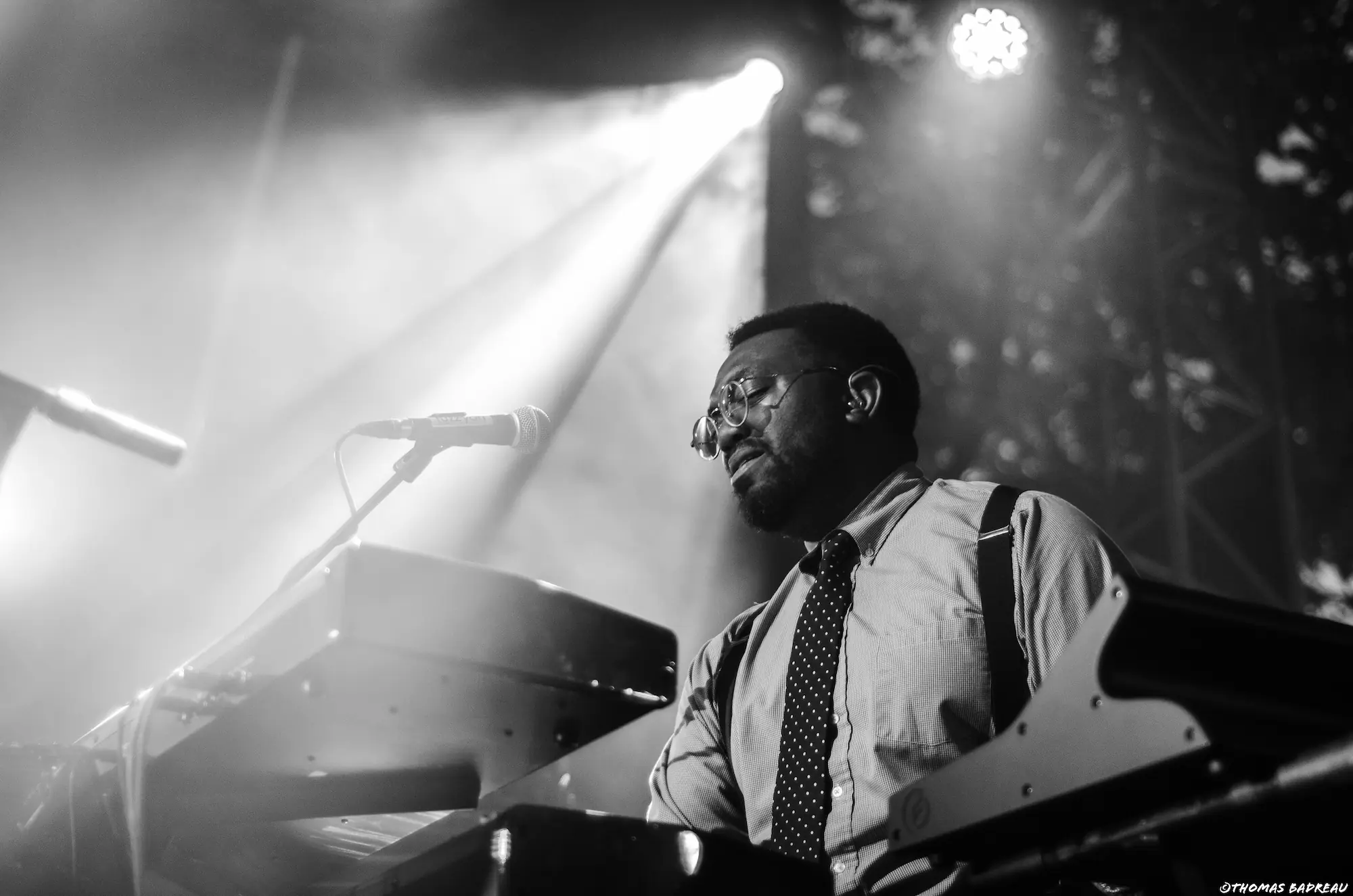concert_8 Un musicien joue du clavier sur scène. Il porte une chemise, une cravate à pois et des bretelles. Il a les yeux fermés, concentré, avec une expression émotionnelle. Une lumière blanche intense éclaire la scène derrière lui, créant une atmosphère dramatique.