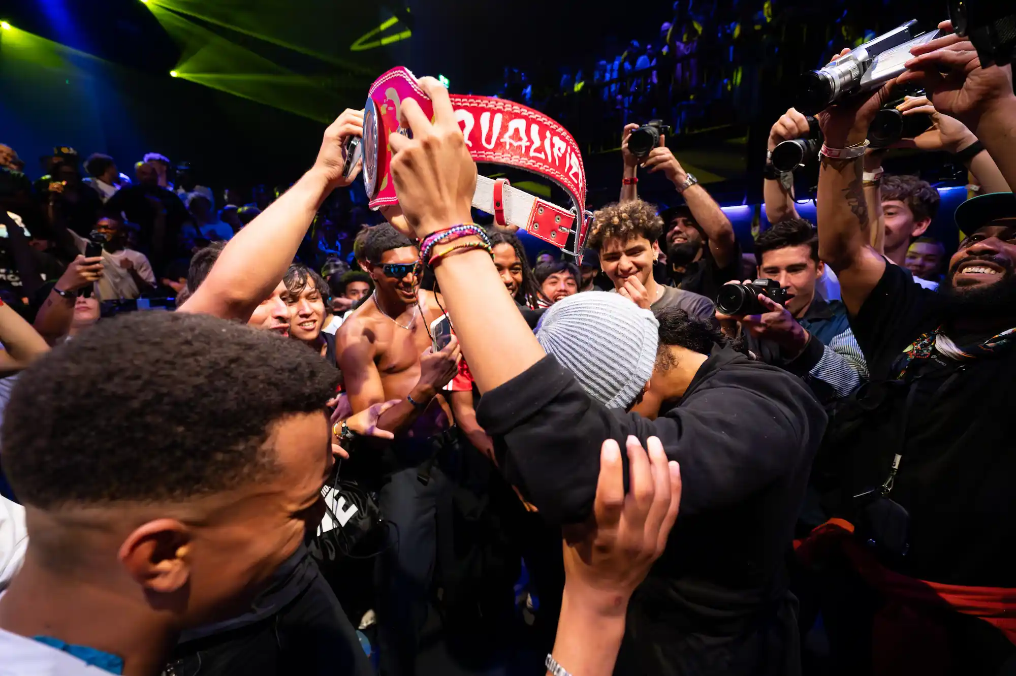 Un groupe de personnes célèbrent dans une ambiance festive et animée. Au centre, une personne portant un bonnet gris lève un large ceinturon rouge marqué « QUALIFIER ». Autour d’eux, plusieurs spectateurs sourient, prennent des photos et filment avec des caméras. L’éclairage est coloré, avec des projecteurs jaunes et bleus créant une atmosphère de fête.