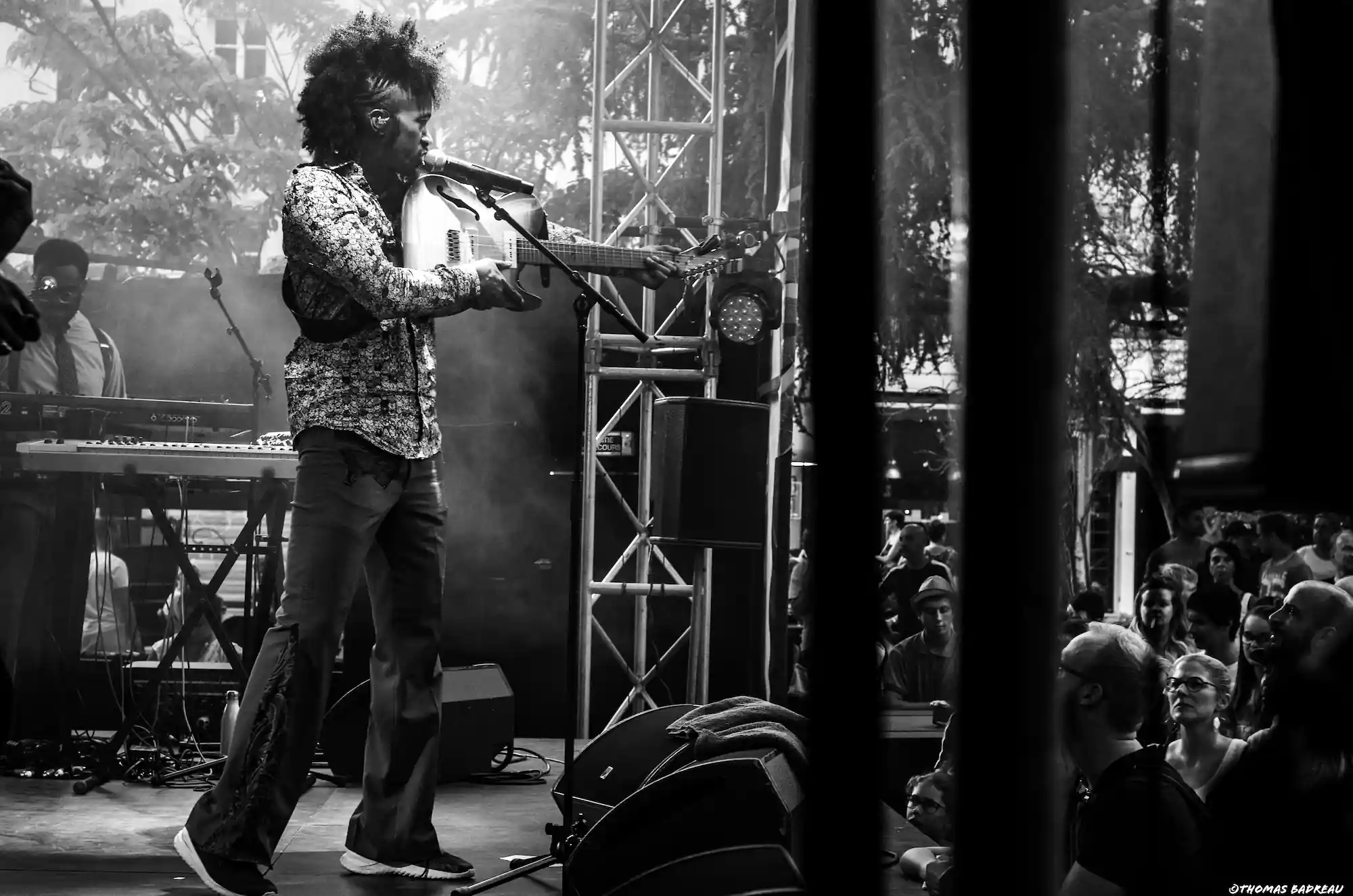 Un musicien joue de la guitare sur scène, chantant dans un micro. Il porte une chemise à motifs et un pantalon. En arrière-plan, un autre musicien est visible près d’un clavier. Le public regarde la performance avec attention. L’image est en noir et blanc.