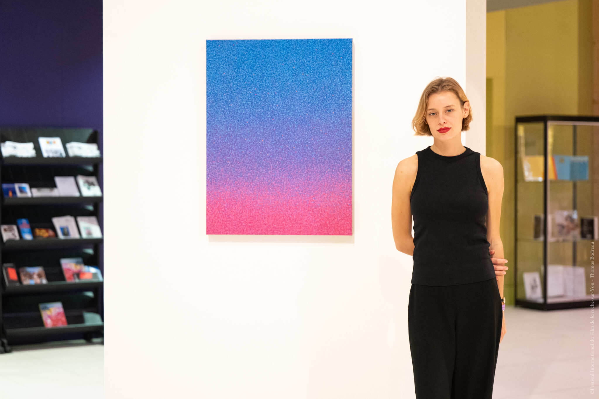 Une femme aux cheveux courts et roux clair, vêtue d’un haut sans manches noir et d’un pantalon noir, pose debout près d’un mur blanc sur lequel est accroché un tableau aux couleurs dégradées du bleu en haut au rose en bas. En arrière-plan, une étagère avec des livres et un meuble vitré sont visibles.