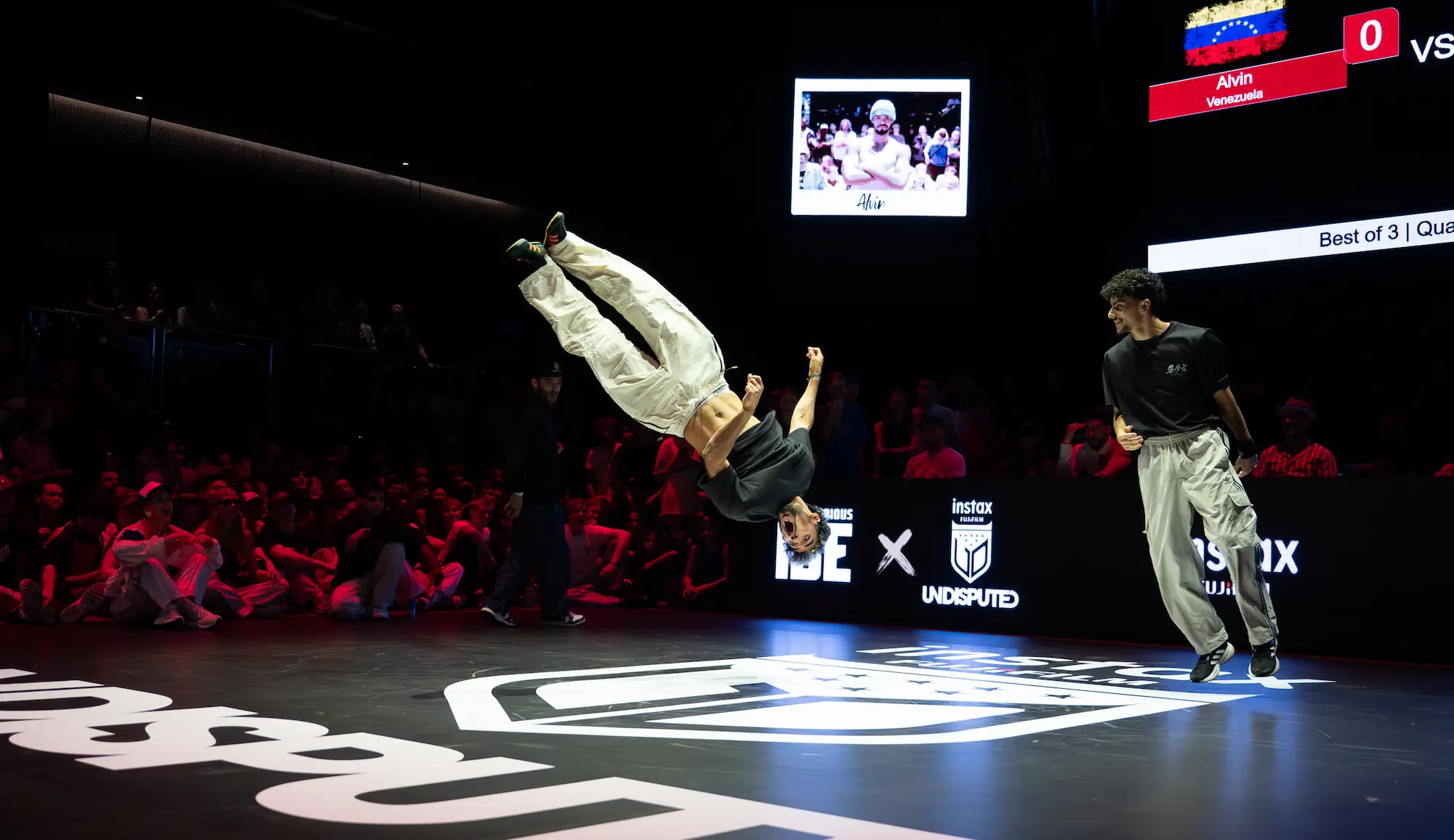 Un danseur de breakdance effectue un salto arrière en l'air, le corps tendu et tête en bas, portant un t-shirt noir et un pantalon blanc large. Un autre danseur, souriant, est debout à droite, vêtu d'un t-shirt noir et d'un pantalon gris clair. En arrière-plan, un public nombreux observe la scène, avec des éclairages sombres et des panneaux lumineux affichant des logos, dont "instax FUJIFILM" et "THE NOTORIOUS IBE x UNDISPUTED". En haut à droite, un écran indique un duel entre "Alvin" du Venezuela et un adversaire inconnu.