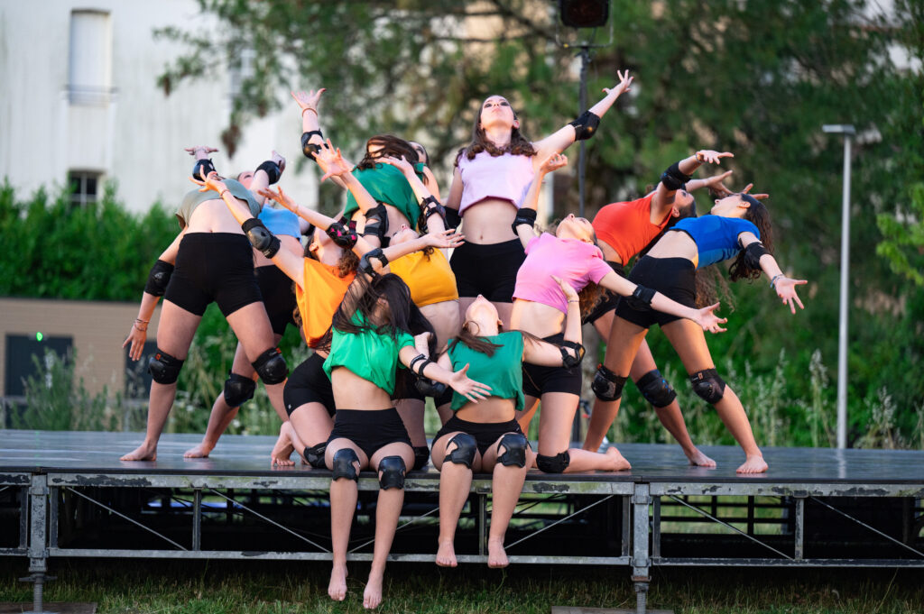 Un groupe de dix danseuses en plein air, sur une scène surélevée. Elles portent des hauts colorés et des shorts noirs, ainsi que des genouillères noires. Elles sont dans une pose expressive et dynamique, avec les bras levés et les têtes rejetées en arrière, formant une composition chorégraphique en groupe. L’arrière-plan montre de la verdure et des bâtiments flous.