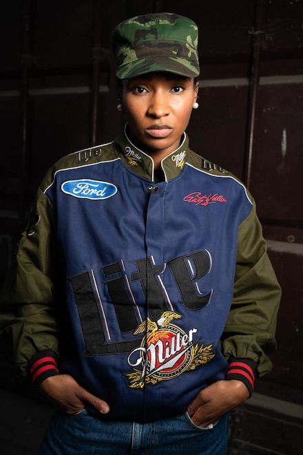 Femme portant une casquette camouflage et une veste de style racing aux logos Ford, Miller Lite et Coca-Cola, posant les mains dans les poches devant un fond sombre.