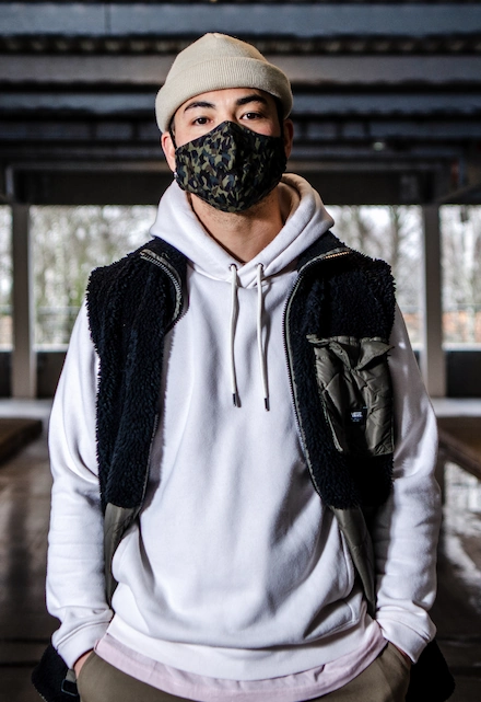 Homme portant un bonnet beige, un masque de protection camouflage, un sweat à capuche blanc et une veste sans manches en polaire noire avec une poche kaki, posant dans un parking couvert.