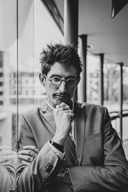 Homme au style affirmé posé contre une vitre, portant un costume clair, des lunettes rondes, plusieurs bagues et bracelets. Il regarde la caméra avec une expression sérieuse, la main posée contre son menton, dans un couloir moderne vitré.