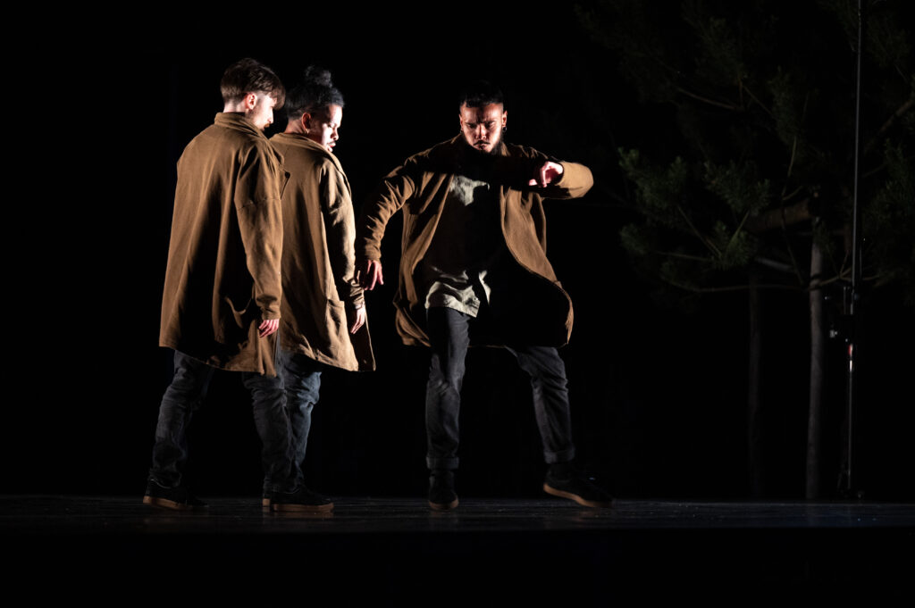 Trois danseurs masculins portent des manteaux marron et des pantalons sombres. Ils sont sur scène, éclairés par une lumière intense qui crée un fort contraste avec le fond noir. L’un des danseurs est en mouvement, levant un pied avec intensité, tandis que les deux autres sont immobiles à ses côtés.