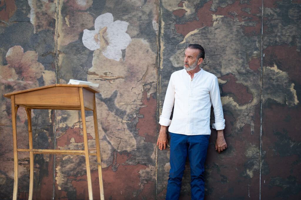 Un homme d’âge mûr, habillé d’une chemise blanche et d’un pantalon bleu, se tient debout, appuyé contre un mur peint d’un motif floral vieilli et délavé. À sa gauche, un petit bureau en bois clair avec un livre posé dessus. L’homme regarde vers la droite, l’air pensif.