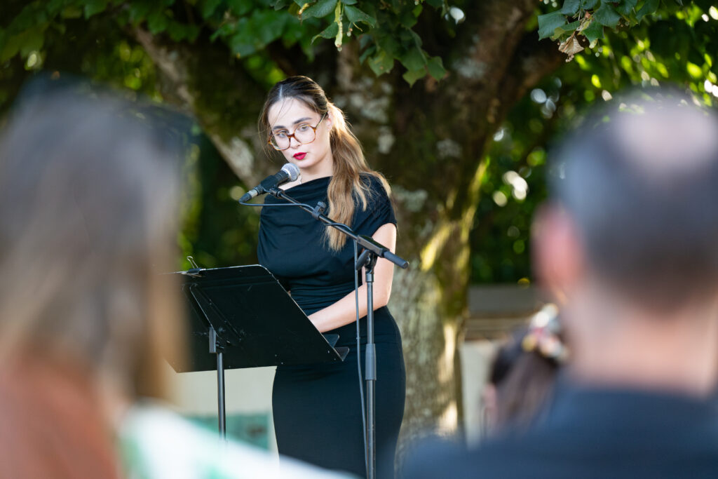 au premier plan en flous il y a 2 têtes de part et d'autre de l'image laissant entrevoir au centre une femme habillé en robe noir sous un arbre et devant un pupitre parlant au micro