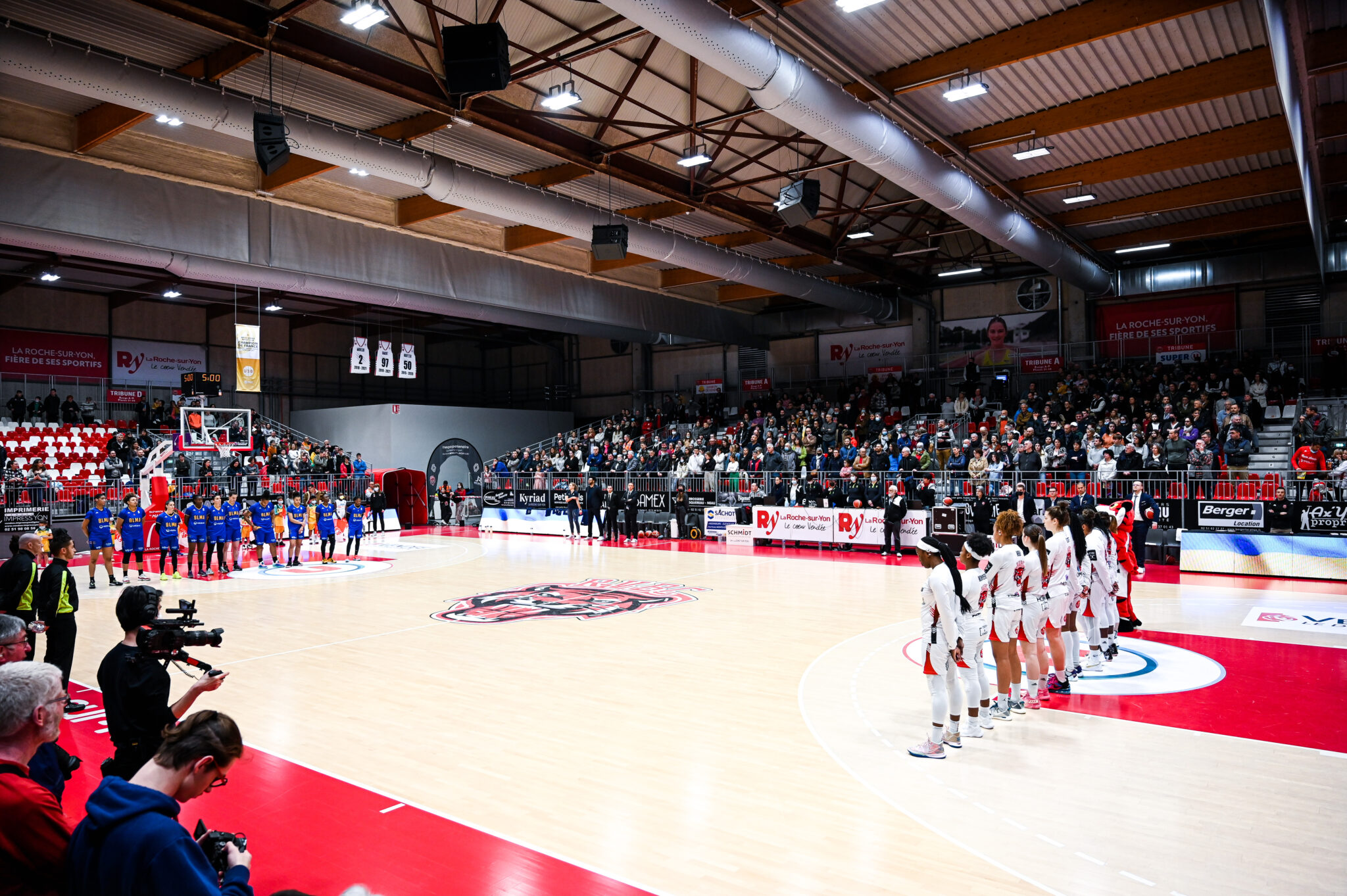 Deux équipes féminines de basketball alignées au centre du terrain pour la présentation avant le début du match. Les joueuses en bleu sont à gauche, celles en blanc à droite. Le public est nombreux dans les gradins d’un gymnase bien éclairé.