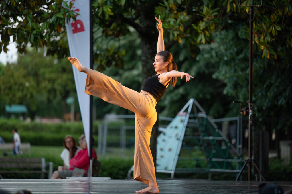 Une danseuse en plein air exécute un mouvement gracieux, une jambe levée à l'horizontale. Elle porte un pantalon ample beige et un débardeur noir, ses bras sont étendus avec élégance. Le décor en arrière-plan montre des arbres et quelques personnes assises, observant la performance.