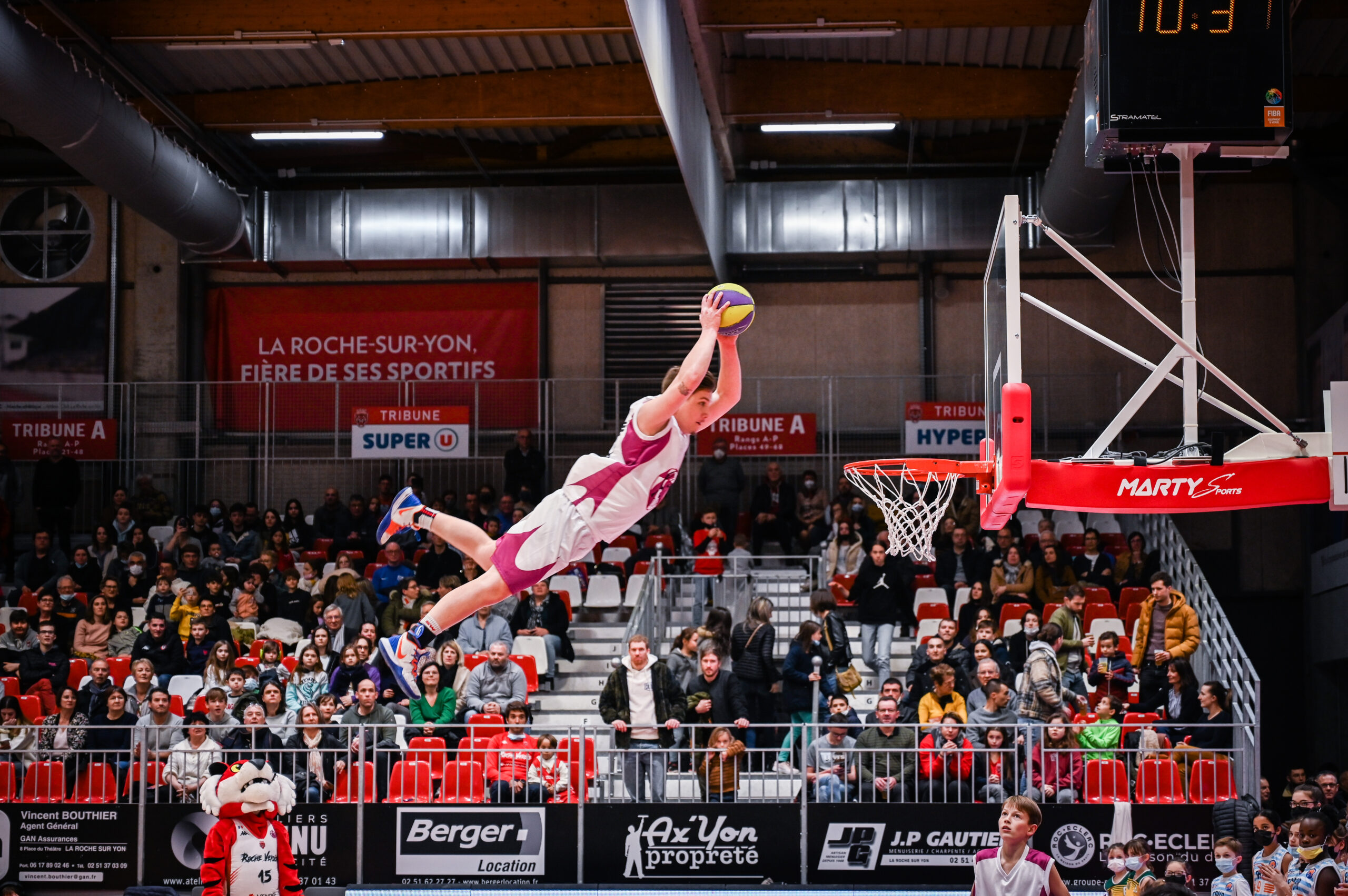 En intérieur, un joueur de basketball en maillot blanc et rose est en suspension dans les airs, tenant le ballon à deux mains au-dessus de sa tête, en direction du panier. Le public est attentif dans les gradins rouges et blancs. Une mascotte est visible en bas à gauche.