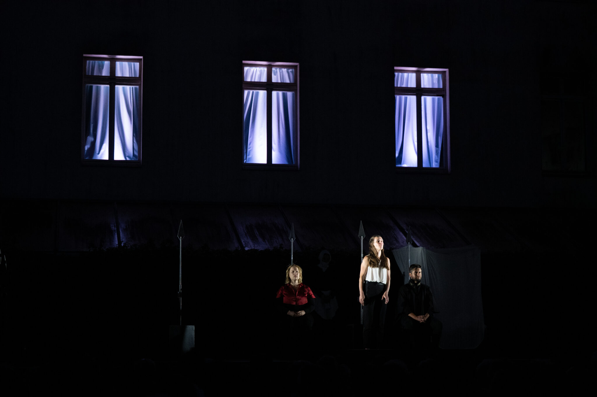 Scène de théâtre en extérieur, de nuit. Trois personnes sont sur scène devant une façade de bâtiment avec trois fenêtres éclairées et drapées. Deux personnes sont assises, une au centre à gauche en chemise rouge et l’autre à droite en tenue sombre, tandis qu’une femme debout au centre porte un haut blanc et un pantalon noir. La scène est peu éclairée, avec une ambiance dramatique.
