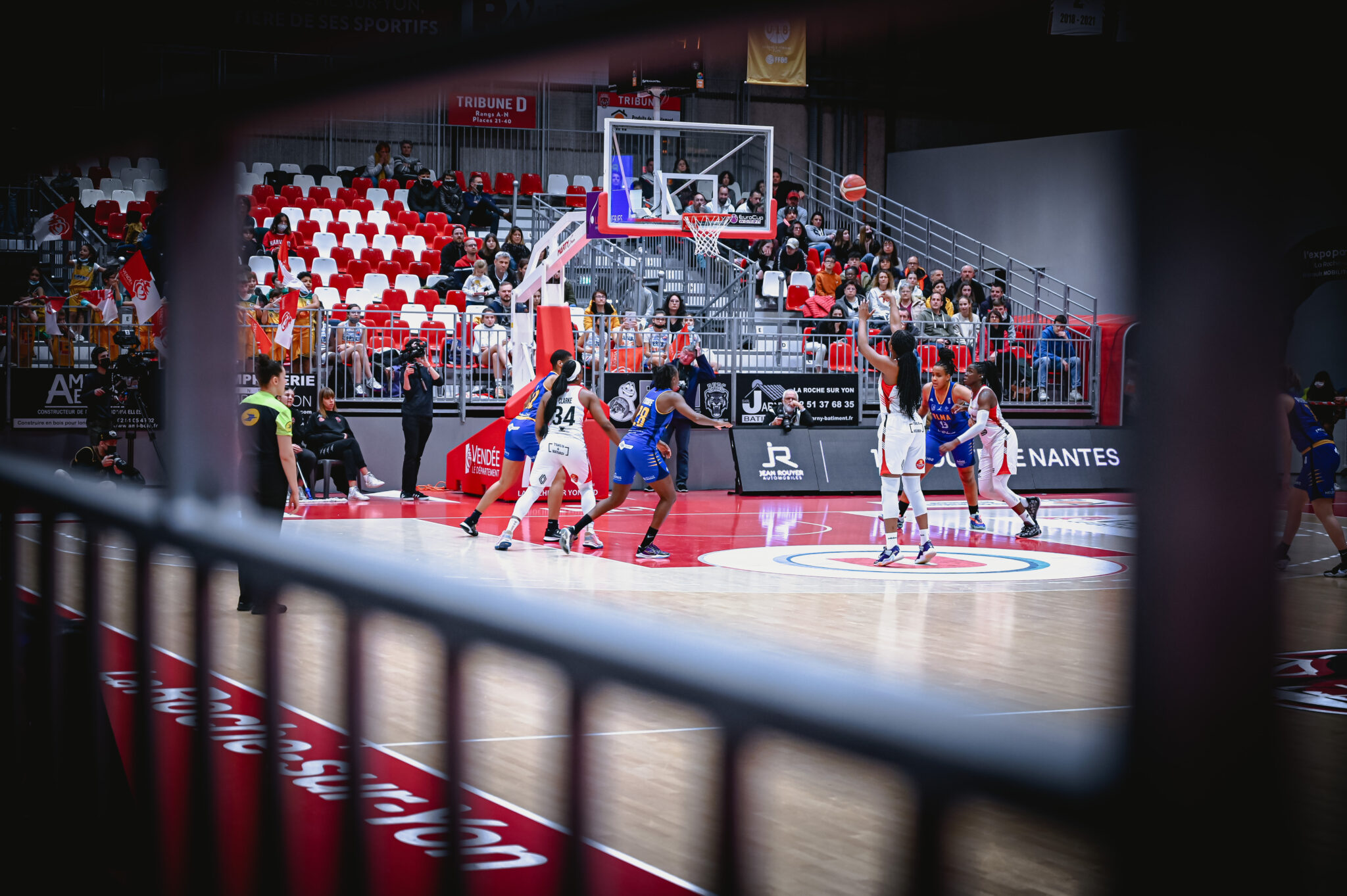 Match de basketball féminin en salle. Une joueuse en blanc vient de tirer au panier, le ballon est en l’air près du panier. Plusieurs joueuses en blanc et bleu sont positionnées sous le panier prêtes à récupérer le ballon. Le public est attentif. La photo est prise à travers une grille.