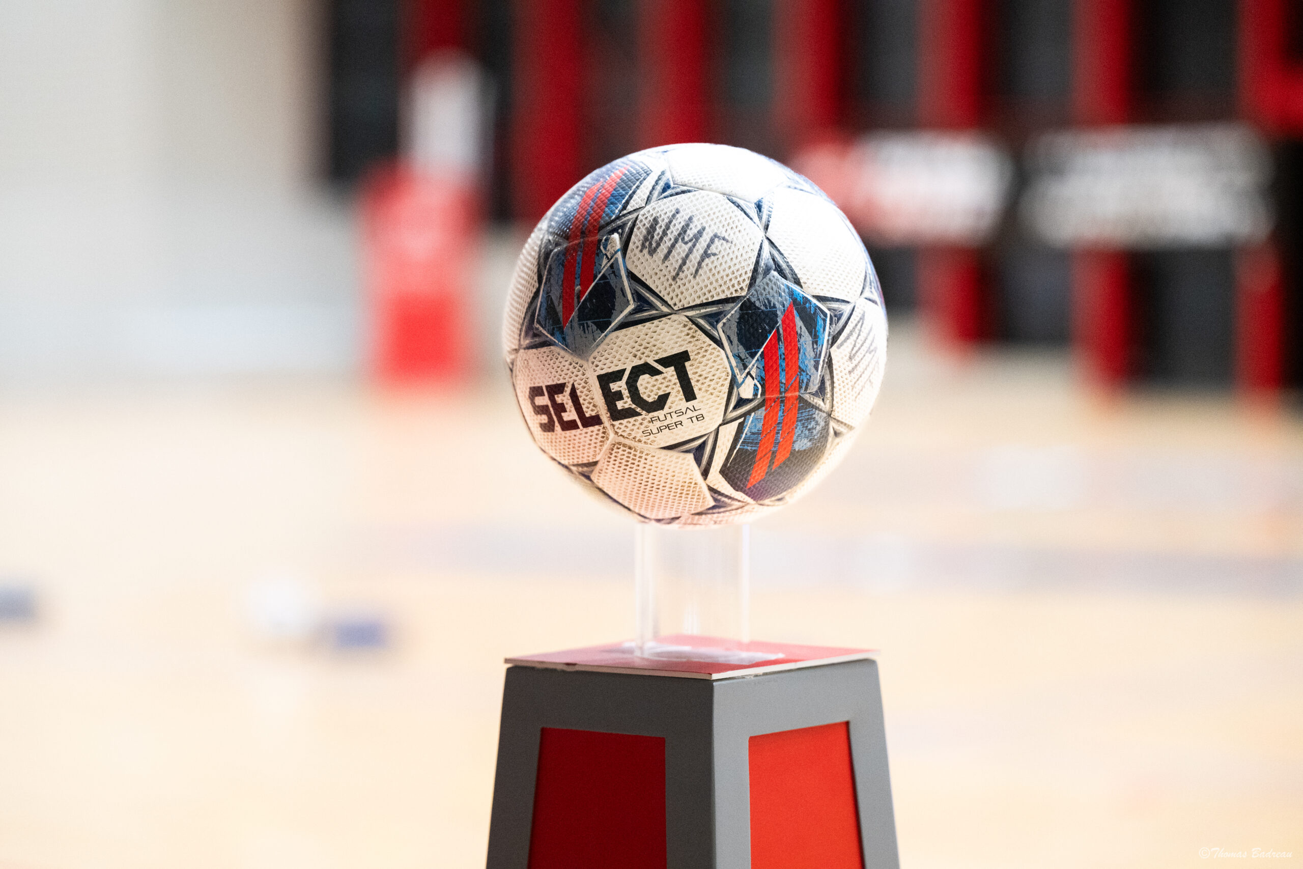 Un ballon de futsal Select Futsal Super TB posé sur un socle rouge et gris, prêt pour le coup d’envoi sur un terrain en salle.