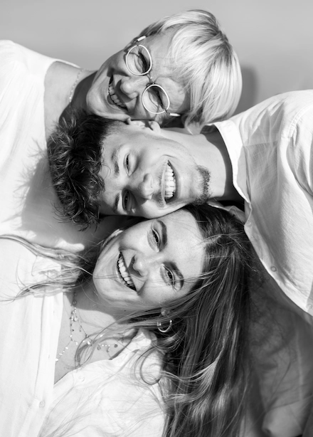 Trois personnes souriantes posent ensemble pour une photo en noir et blanc. Une femme plus âgée avec des lunettes est en haut, un jeune homme au centre repose sa tête sur l’épaule d’une jeune femme en bas. Tous trois sont proches, joyeux, et regardent l’objectif.