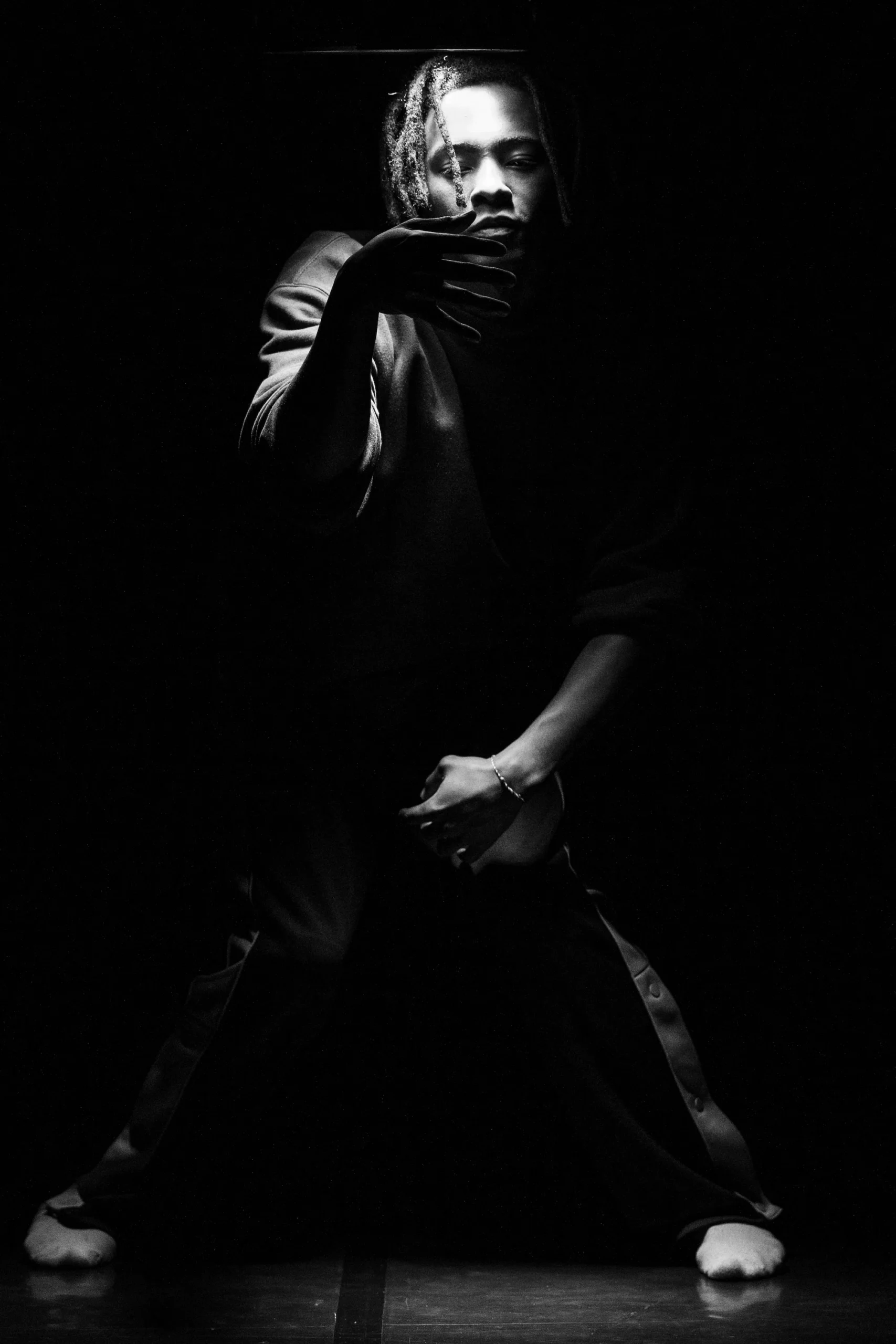 Un portrait en noir et blanc d'une personne debout dans une pose artistique, partiellement éclairée par une lumière dramatique. Elle est pieds nus, avec les jambes écartées et une main placée devant son visage, créant un effet d’ombre saisissant. Ses cheveux tombent en tresses sur son front. L’image évoque une ambiance introspective et expressive, accentuée par le contraste fort entre lumière et obscurité.