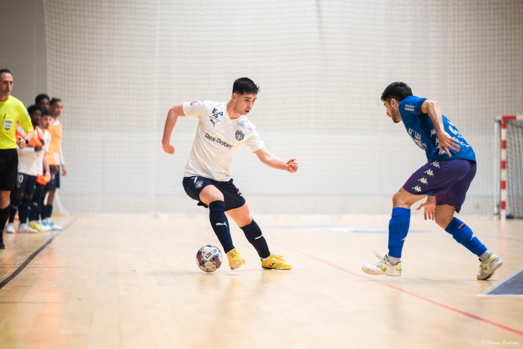 Un joueur de futsal en maillot blanc tente de dribbler un adversaire en maillot bleu sur un terrain intérieur, sous le regard de l’arbitre et des remplaçants alignés sur le côté.