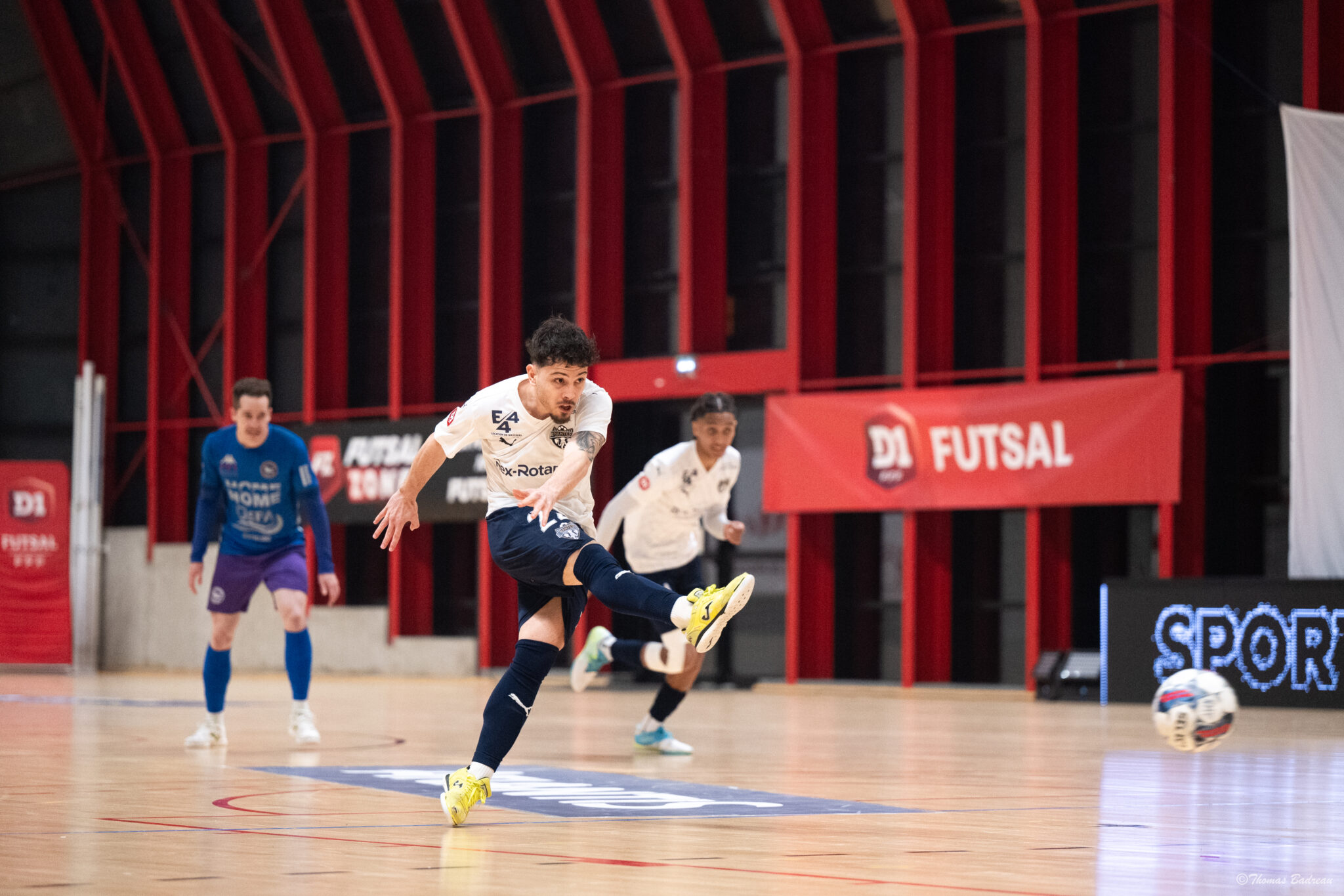 Un joueur de futsal en maillot blanc frappe le ballon en pleine action sur un terrain indoor, sous les regards d'autres joueurs. Une bannière rouge ‘D1 FUTSAL’ est visible en arrière-plan.