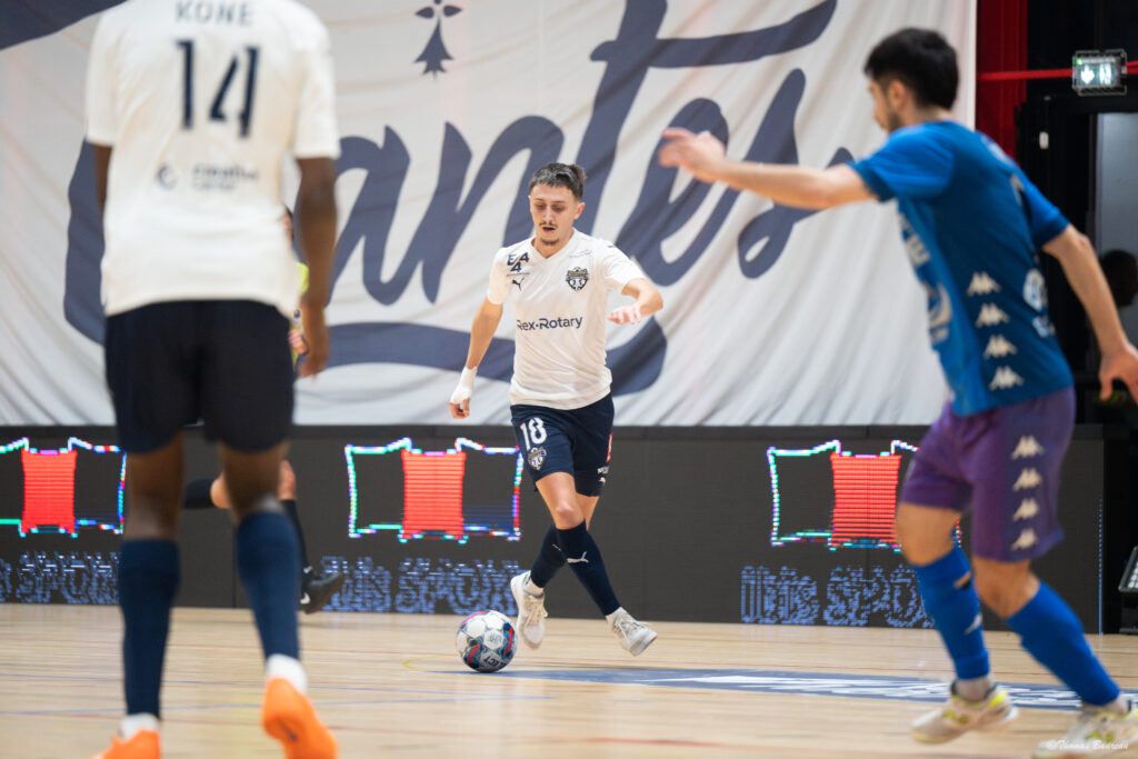Un joueur de futsal en maillot blanc, ballon au pied, avance sur le terrain face à des adversaires en bleu. Une grande bannière marquée ‘Nantes’ est visible en arrière-plan.
