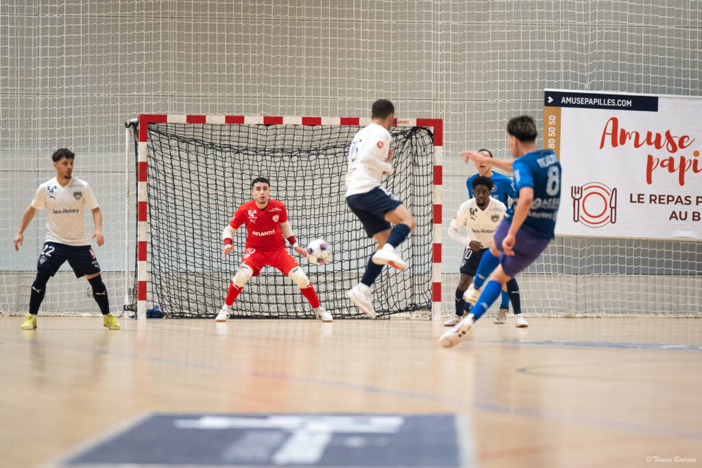 Une action de futsal intense devant le but : un joueur en bleu frappe le ballon tandis que le gardien en rouge, concentré, se prépare à bloquer le tir. Plusieurs joueurs en blanc défendent devant le filet, avec une publicité ‘Amuse papilles’ visible en arrière-plan.