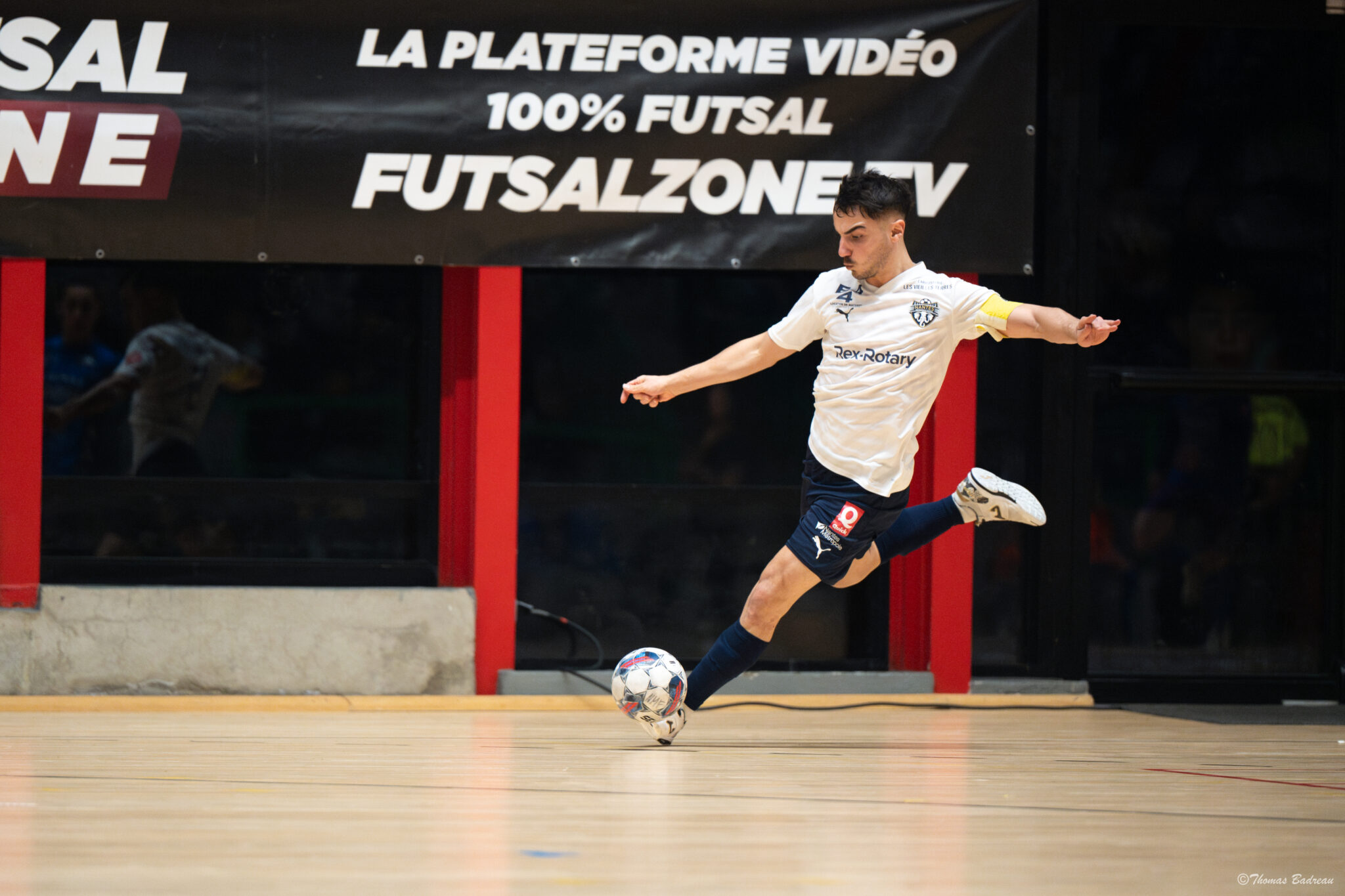 Un joueur de futsal en plein tir frappe le ballon du pied droit sur un terrain intérieur. Il porte un maillot blanc et un short bleu marine, avec une banderole en arrière-plan indiquant ‘Futsalzone.tv – La plateforme vidéo 100% futsal’.