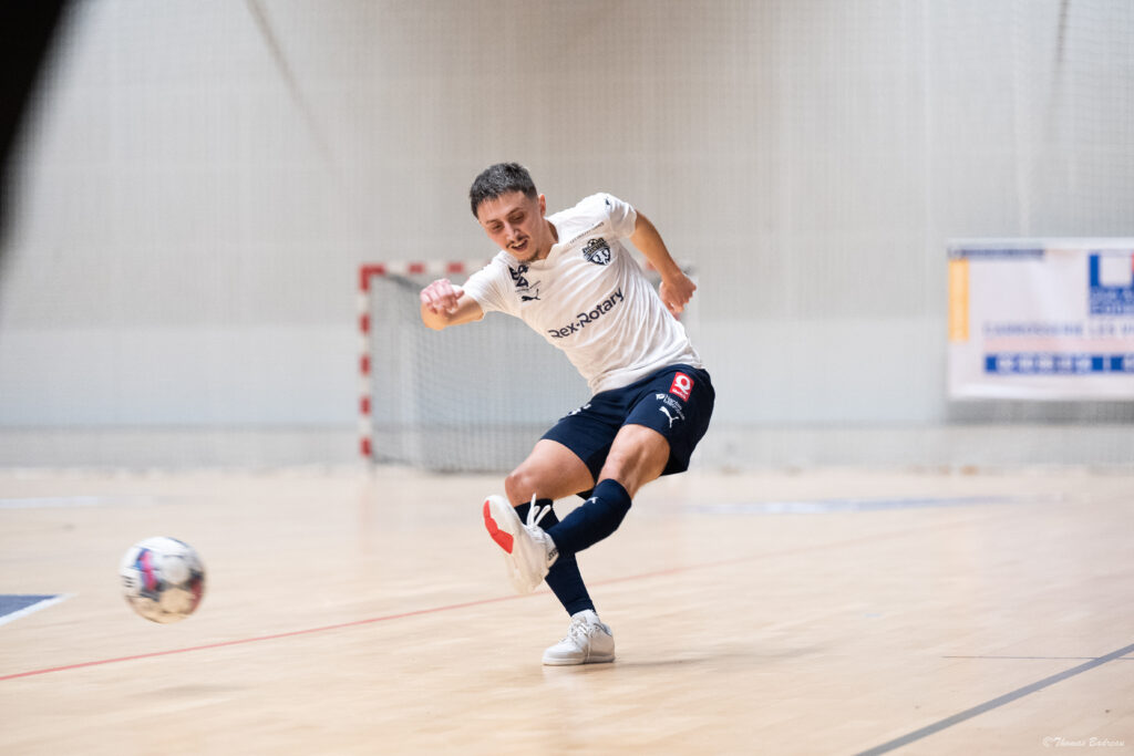 Un joueur de futsal en pleine action frappe le ballon sur un terrain intérieur. Il porte un maillot blanc, un short bleu marine et des chaussures de sport blanches. En arrière-plan, un but et une affiche sont visibles.