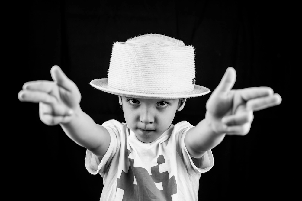 Photo en noir et blanc d’un enfant portant un chapeau clair et un t-shirt, faisant un geste de mains imitant des pistolets vers l’avant, regardant intensément l’objectif sur un fond noir.