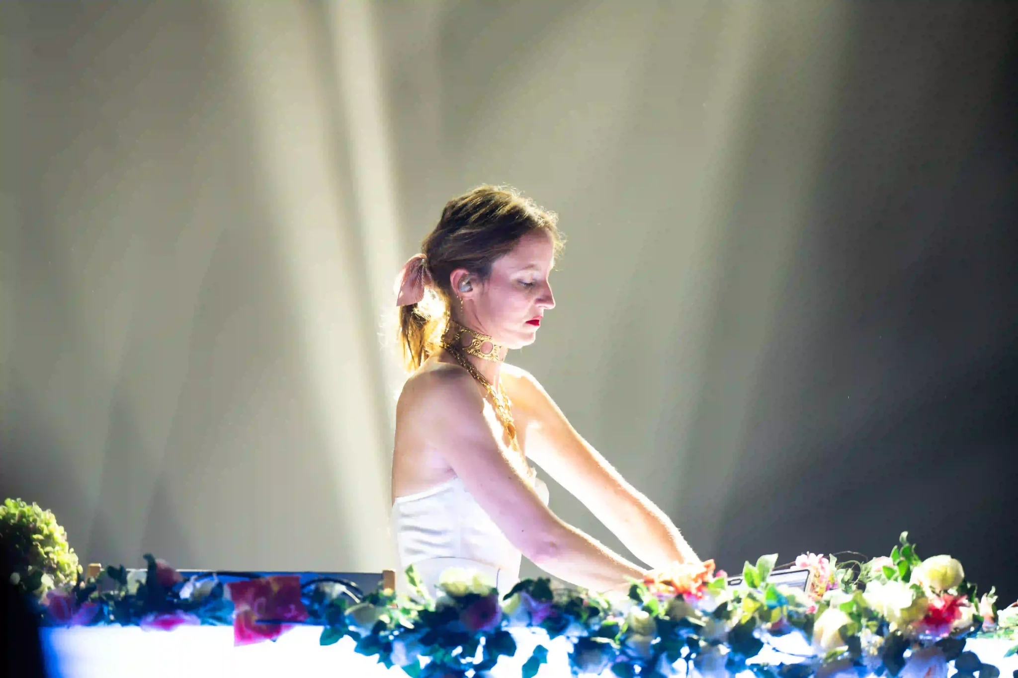 dresel_concert_5 Une DJ féminine au visage concentré est éclairée par une lumière blanche intense. Elle porte une robe blanche, un choker doré et une queue de cheval attachée avec un ruban rose. Devant elle, une console lumineuse décorée de fleurs colorées.