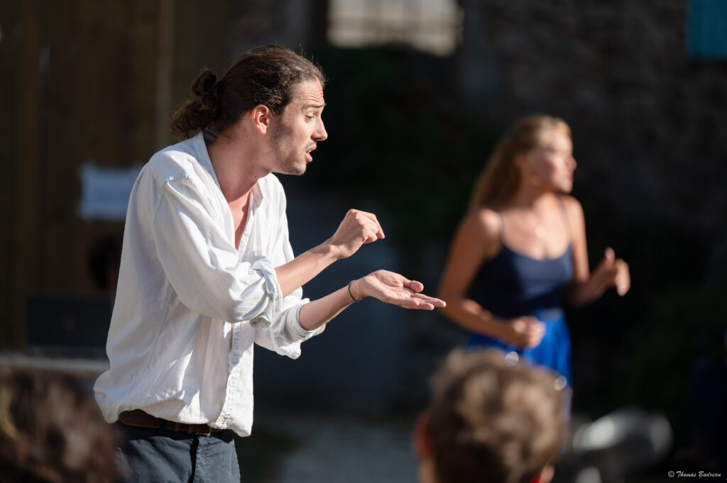 Un homme à cheveux longs attachés en queue de cheval, portant une chemise blanche, s’exprime avec intensité en utilisant des gestes de ses mains. En arrière-plan, une femme floue en débardeur noir et short bleu semble aussi en train de parler ou d’agir. La scène se déroule en extérieur, avec un fond sombre et des spectateurs visibles au premier plan.