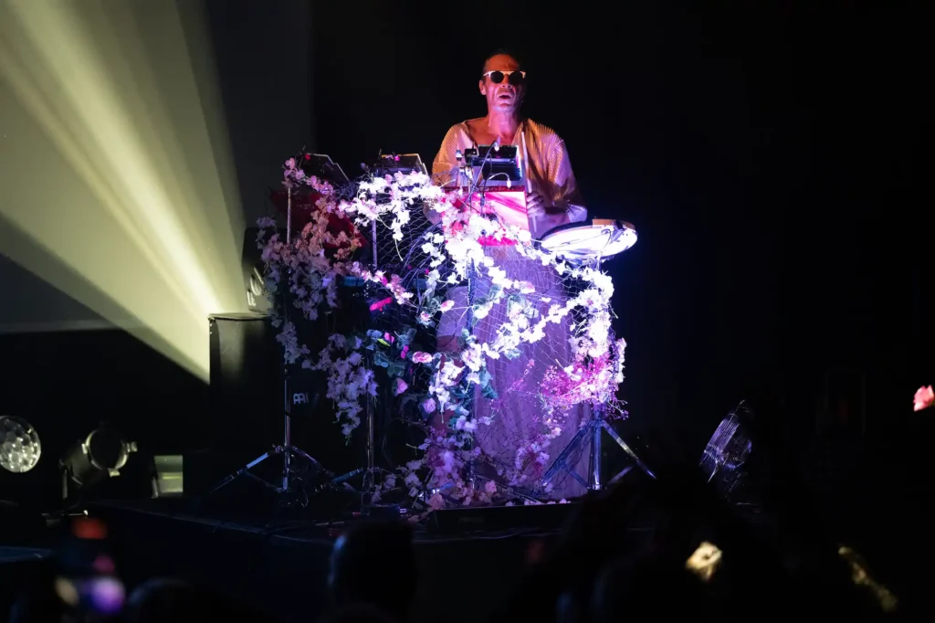 Un DJ portant des lunettes de soleil et une tenue ample claire se tient derrière une console ornée de guirlandes de fleurs blanches. La scène est plongée dans une lumière sombre avec un projecteur jaune derrière lui, créant une atmosphère intime et mystérieuse.