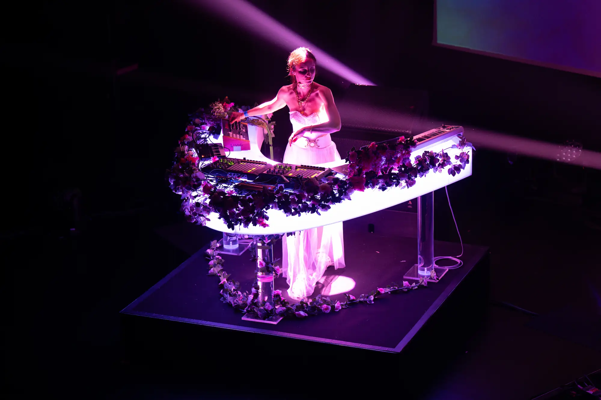 dresel_concert_3 Une artiste DJ en robe blanche élégante est sur scène, illuminée par une lumière violette. Elle est derrière une console en forme de demi-cercle, décorée de guirlandes de fleurs. La console est équipée de plusieurs appareils électroniques et claviers. L'ambiance est intimiste avec un fond sombre.