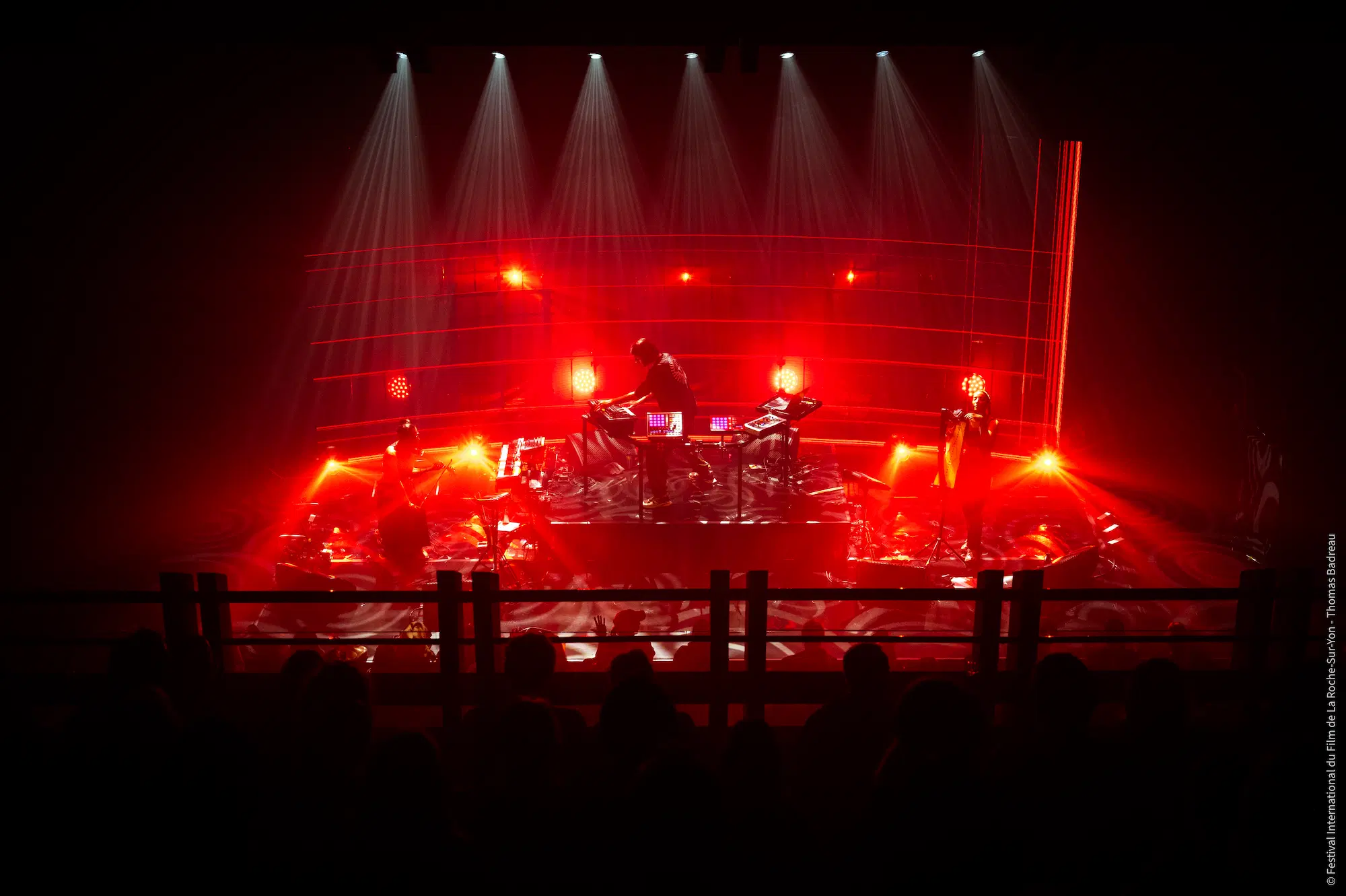 Scène de concert vue de haut avec un éclairage rouge intense. Au centre, un musicien est debout derrière une table équipée de plusieurs claviers et contrôleurs électroniques. Deux autres musiciens sont présents sur scène, jouant d'instruments différents. Des faisceaux de lumière blanche descendent du plafond, illuminant la scène et créant une ambiance dramatique. Au premier plan, l'ombre du public est visible.
