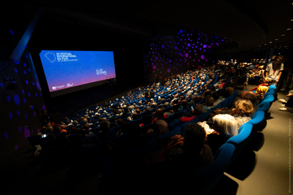 salle de spectacle avec une tribune siège bleu et une scène noir avec grand écran