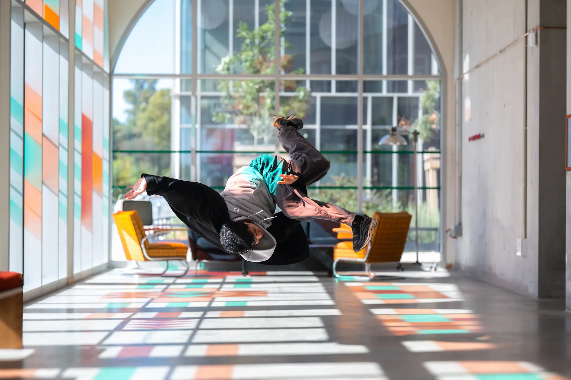 Un danseur en plein saut acrobatique à l’intérieur d’une pièce lumineuse avec de grandes fenêtres et un sol aux motifs colorés. Il porte un sweat à capuche multicolore, un pantalon foncé et des baskets noires. En arrière-plan, on aperçoit un canapé noir et deux fauteuils orange.