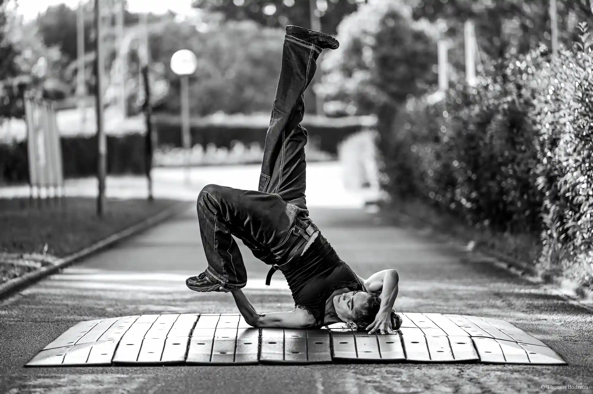 En noir et blanc, un danseur de breakdance exécute un mouvement acrobatique d’équilibre sur un bras, sur un tapis posé au sol, dans un environnement urbain extérieur. Il porte un débardeur noir, un pantalon ample et des chaussures foncées. Son autre bras est plié et sa main touche sa tête. Le fond montre une allée bordée d’arbustes et des panneaux flous.
