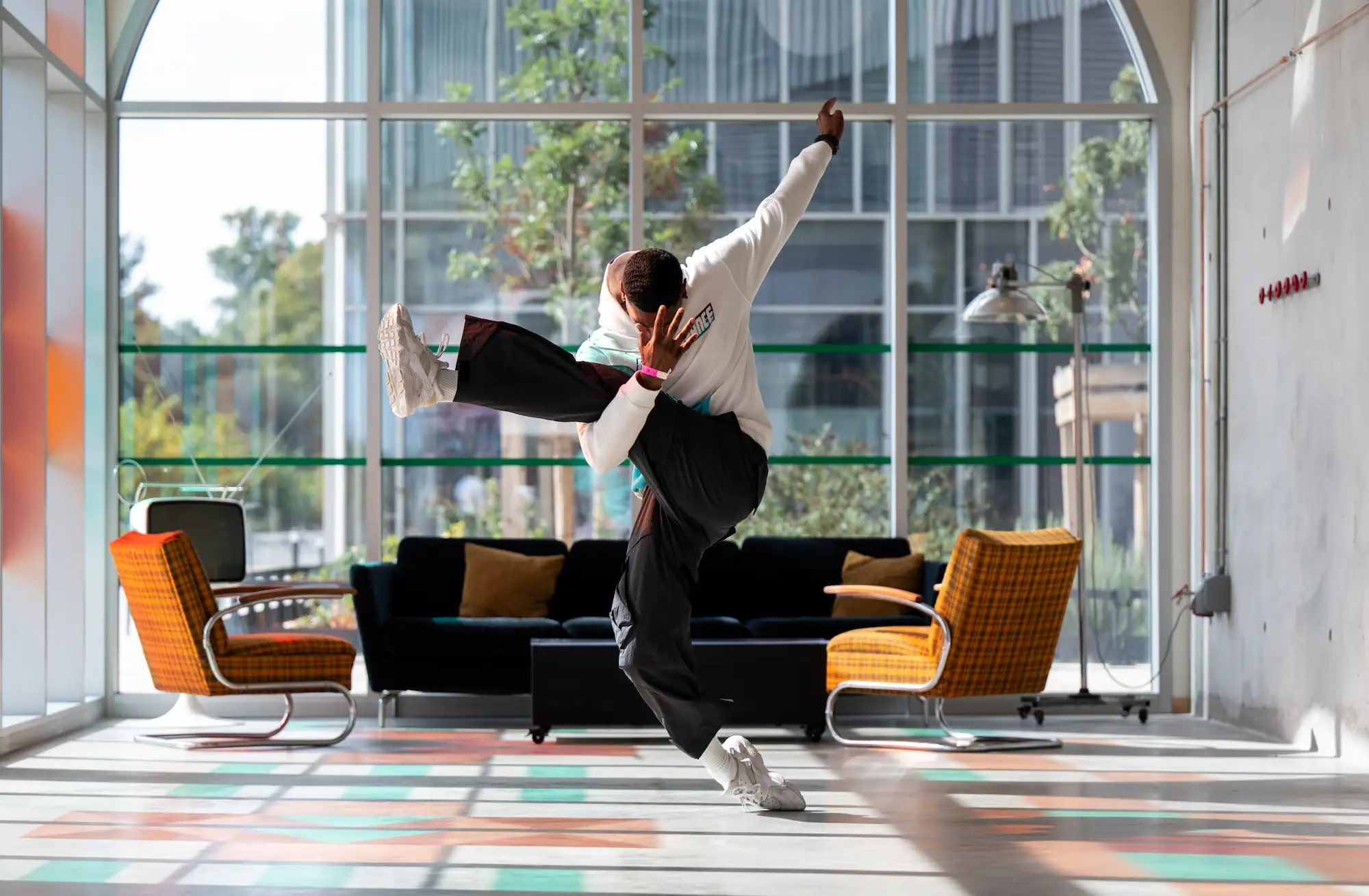 Un danseur urbain réalise un mouvement dynamique de danse en équilibre sur une jambe, dans un intérieur lumineux avec de grandes fenêtres. Il porte un sweat blanc, un pantalon noir large, des baskets blanches, et une montre noire. Le décor comprend deux fauteuils orange et un canapé noir avec des coussins, ainsi qu’une vieille télévision et une lampe sur pied.