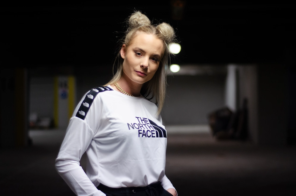 Jeune femme debout dans un parking sombre, portant un sweat blanc 'The North Face' avec des bandes logo sur les épaules. Elle a les cheveux blonds coiffés en deux petits chignons et regarde calmement l’objectif, une main dans la poche.