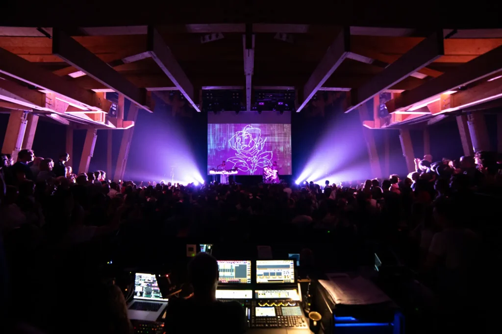 derrière la régis nous voyons une salle de spectacle avec une DJ sur scène avec écran géant et jeux de lumière violet
