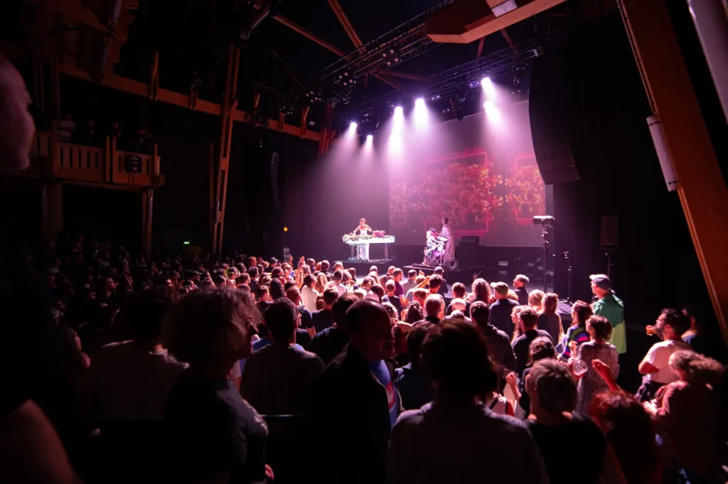 Une foule nombreuse assiste à un concert en intérieur. Sur scène, deux artistes sont éclairés par des projecteurs violets. L'un des artistes est derrière un pupitre décoré de fleurs, tandis que l'autre semble manipuler un dispositif décoratif floral. En arrière-plan, un grand écran projette des images colorées. La salle est sombre autour de la scène, avec une structure en bois visible sur les côtés.
