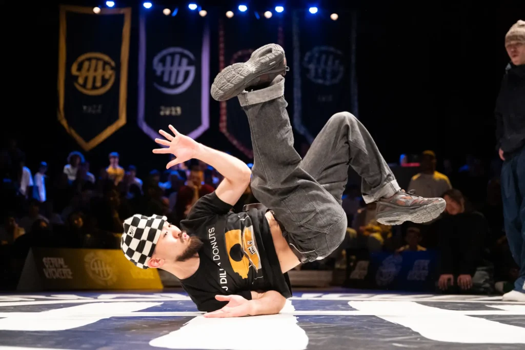 Un danseur de breakdance réalise un mouvement au sol en appui sur un avant-bras, avec les jambes levées et pliées. Il porte un t-shirt noir avec un motif jaune et blanc, un pantalon gris et des chaussures de sport. Sur la tête, un bonnet à damiers noir et blanc. En arrière-plan, un public nombreux observe la performance dans une salle sombre avec des bannières accrochées.