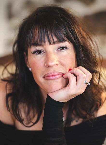 Portrait d’une femme aux cheveux noirs avec frange, posant la main sous le menton, regardant l’objectif avec un léger sourire.