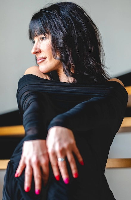 L’image montre une femme vêtue d’une robe noire à manches longues, posant de manière gracieuse avec les bras croisés sur les genoux. Elle regarde vers la gauche avec un léger sourire. Ses cheveux sont foncés, mi-longs et ondulés, avec une frange couvrant partiellement son front. Elle porte du vernis rouge-rose vif sur les ongles et une bague argentée à l’annulaire gauche. L’arrière-plan est flou, avec des éléments en bois et mur blanc, suggérant un intérieur moderne, peut-être un escalier. La lumière naturelle éclaire son visage, donnant à la scène une ambiance douce et intime.